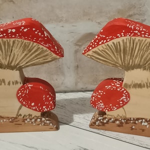 May include: Two wooden mushroom figurines with red and white caps and brown stems. The mushrooms are painted with white dots and have a textured surface.