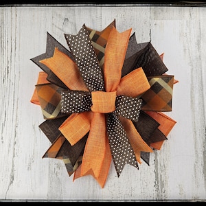 May include: A decorative bow featuring layers of orange, brown, and plaid ribbons. The bow has a central knot and is arranged in a circular pattern. The ribbons have polka dot and plaid patterns.