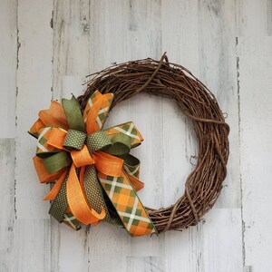 May include: A grapevine wreath with a large bow made of orange and green plaid ribbon. The bow is tied with a green ribbon with white polka dots.