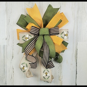 May include: A green, yellow, and brown bow with a bee design. The bow is made of burlap and ribbon and is tied with a wire.