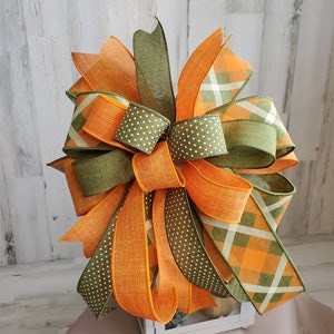 May include: A large, multi-layered bow made with orange, green, and white ribbon. The bow is tied with a plaid ribbon and a polka dot ribbon. The bow is sitting on top of a white lantern.