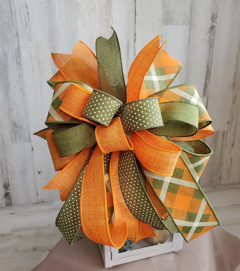 May include: A large, multi-layered bow made with orange, green, and white ribbon. The bow is tied with a plaid ribbon and a polka dot ribbon. The bow is sitting on top of a white lantern.