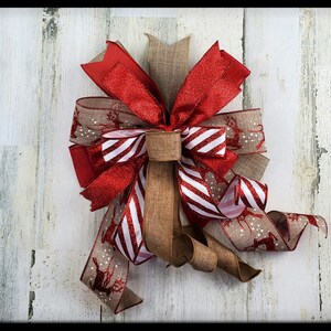 May include: A decorative Christmas bow featuring red, white, and brown ribbons. The bow has a red glitter ribbon, a striped red and white ribbon, and a brown ribbon with reindeer and polka dots. The bow is set against a white wooden background.