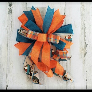May include: A large orange and teal bow with a plaid ribbon and a white ribbon with a pumpkin and wheelbarrow design. The bow is tied with a loop and is ready to be hung on a door or wreath.