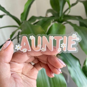 May include: A clear sticker with the word "AUNTIE" in pink letters surrounded by white flowers.
