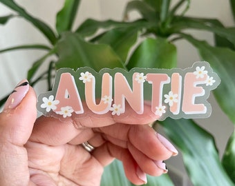 May include: A clear sticker with the word "AUNTIE" in pink letters surrounded by white flowers.