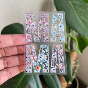 May include: A clear sticker with the word "MAMA" spelled out in large letters. The letters are filled with a floral pattern in shades of pink, blue, and green.