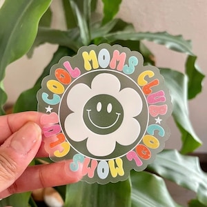 May include: A clear sticker with a white flower with a smiley face in the center. The flower is surrounded by a circle with the text "Cool Moms Club" in rainbow colors.