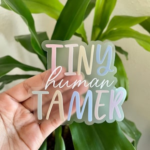 May include: A clear sticker with the text "Tiny human tamer" in colorful, handwritten lettering. The sticker is on a hand, with a green plant in the background.