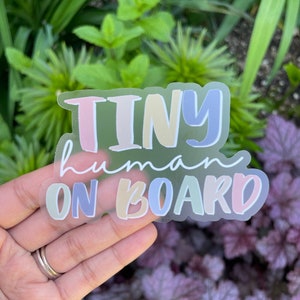 May include: A clear sticker with the text "TINY human ON BOARD" in colorful, handwritten letters. The letters are in shades of pink, yellow, blue, and green.