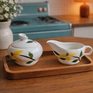 Op de afbeelding: Witte keramische suikerpot met deksel en creamer set op een houten dienblad. De set is versierd met gele bloemen en groene bladeren. De achtergrond omvat een vintage blauw fornuis en een warm getinte lamp.