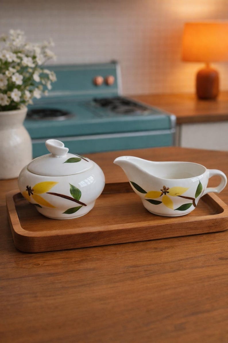 Op de afbeelding: Witte keramische suikerpot met deksel en creamer set op een houten dienblad. De set is versierd met gele bloemen en groene bladeren. De achtergrond omvat een vintage blauw fornuis en een warm getinte lamp.