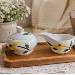 Op de afbeelding: Een witte keramische suikerpot met deksel en een melkkan met gele bloemmotieven en groene bladeren, op een houten dienblad. Een vaas met witte bloemen en een draadmand met eieren staan op de achtergrond.