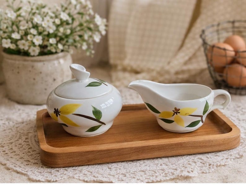 Op de afbeelding: Een witte keramische suikerpot met deksel en een melkkan met gele bloemmotieven en groene bladeren, op een houten dienblad. Een vaas met witte bloemen en een draadmand met eieren staan op de achtergrond.