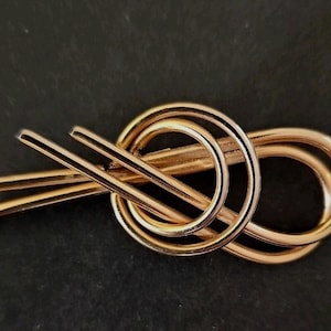 May include: A gold-toned metal brooch with a unique, abstract design featuring intertwined loops and lines.