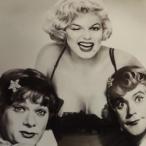 May include: Black and white photograph of three people. A woman with blonde hair winks and smiles, wearing a black top. Two other people with dark hair are in the foreground, one wearing a headband with a flower.