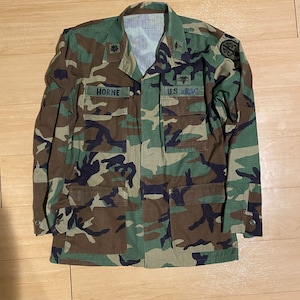 May include: A long-sleeve camouflage field jacket featuring a woodland pattern of green, brown, and black. The jacket includes four front pockets, a collar, and embroidered patches reading HORNE and U.S. ARMY, along with medical insignia.