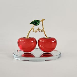 May include: Two red glass cherries with a gold stem and a green leaf, resting on a clear, round mirrored surface. The word "Love" is written in gold on the stem. The cherries are a decorative item.