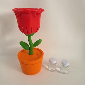 May include: A red rose-shaped bubble machine with a green stem and leaves, set in an orange flower pot base with a small power button. Two small, clear plastic bottles with white caps containing bubble solution are positioned next to the base.