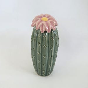 May include: A ceramic cactus-shaped jar with a glossy dark green finish and vertical ridges. The lid is shaped like a pink flower with a yellow center, featuring a smooth, glazed surface that sits atop the textured, spiky-looking cactus body.