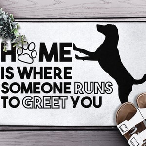 May include: White doormat with black text that reads "Home is where someone runs to greet you". A black silhouette of a dog running is to the right of the text.