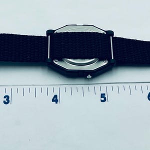 May include: Black watch with a black canvas strap and a silver buckle. The watch face is a dark color and has a unique geometric shape. The watch is shown next to a ruler.