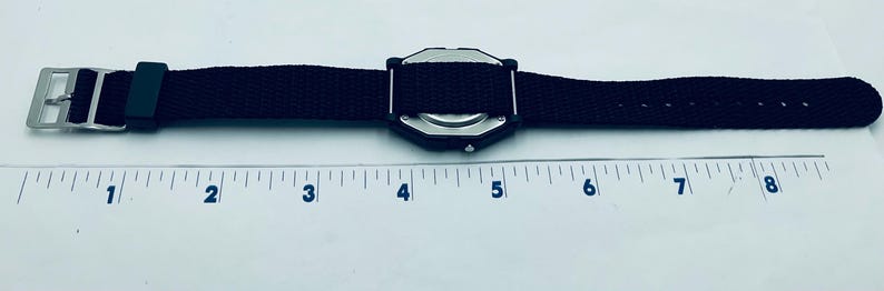 May include: Black watch with a black canvas strap and a silver buckle. The watch face is a dark color and has a unique geometric shape. The watch is shown next to a ruler.