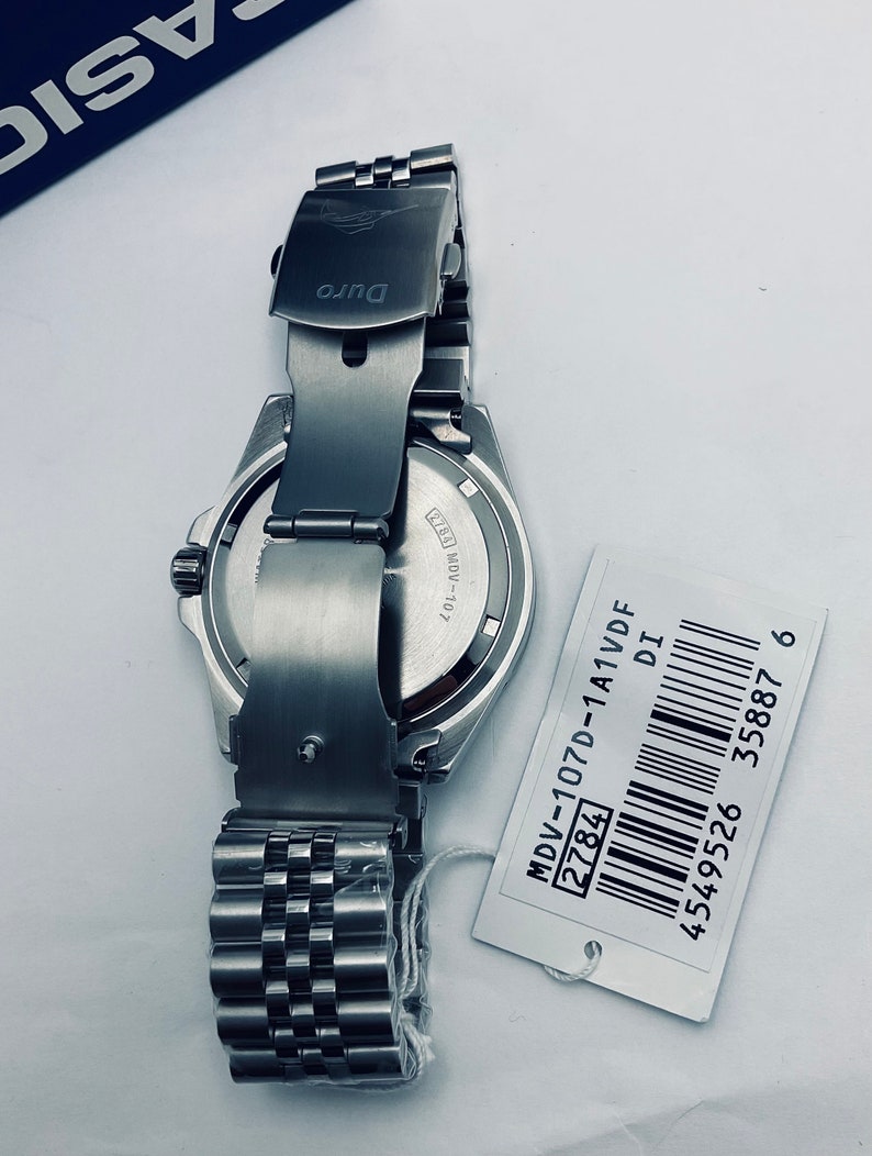May include: A silver watch with a stainless steel strap and a black face. The watch has a date window and a silver clasp. The watch is on a white background with a white tag that says "MDV-107D-1A1VDF 2784 DI 4549526 35887 6".