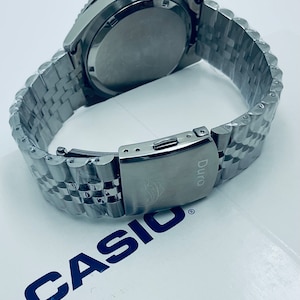 May include: A silver Casio Duro watch with a stainless steel band. The watch has a black face and a silver bezel. The watch is on a white surface with the Casio logo.