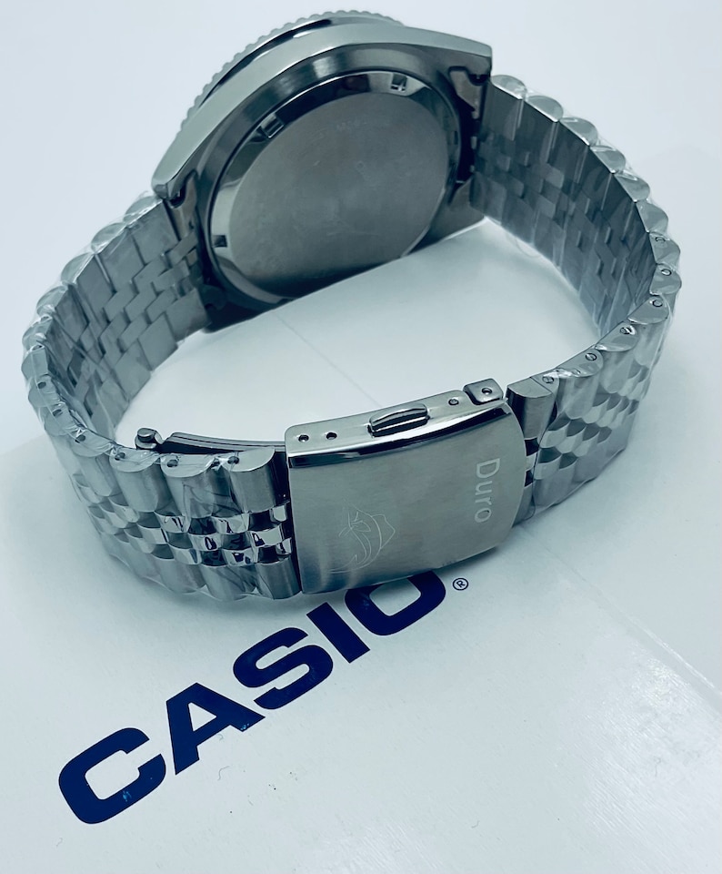 May include: A silver Casio Duro watch with a stainless steel band. The watch has a black face and a silver bezel. The watch is on a white surface with the Casio logo.