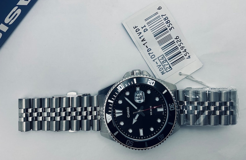 May include: A black and silver Casio watch with a black face and silver hands. The watch has a black bezel with white markings and a silver metal band. The watch is labelled "MDV-107D-1A1VDF" and has a barcode.