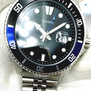 May include: A silver Casio wristwatch with a black face and blue accents. The watch has a silver metal band and a black bezel with white numbers. The watch face has white hands and a date window at the 3 o'clock position. The watch is water resistant to 200 meters.