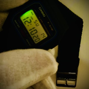 May include: A black Casio digital watch with a black band and a silver buckle. The watch face displays the time, date, and day of the week. The time is 12:20 and the day is Sunday.