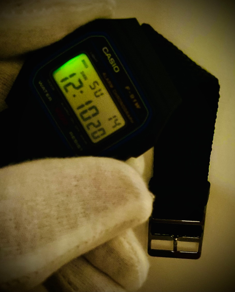 May include: A black Casio digital watch with a black band and a silver buckle. The watch face displays the time, date, and day of the week. The time is 12:20 and the day is Sunday.