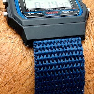 Könnte beinhalten: Eine schwarze Casio F-91W Digitaluhr mit einem blauen Nylonarmband. Das Zifferblatt der Uhr zeigt die Zeit, das Datum und die Alarmeinstellungen an. Die Uhr ist wasserdicht.