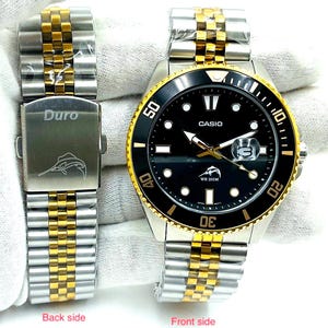 May include: A black and gold Casio watch with a black face and gold hands. The watch has a black bezel with white numbers and a gold band. The watch is water resistant to 200 meters. The back of the watch has the word "Duro" engraved on it.