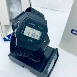May include: A black Casio F-91W digital watch with a black fabric strap. The watch displays the time 4:36:31. The watch is on a black display stand. The watch has the text "CASIO" and "F-91W" on the face.