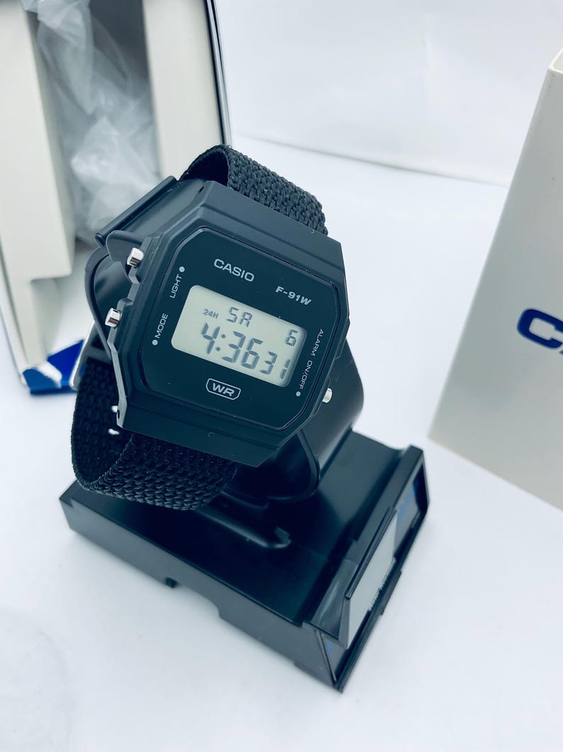 May include: A black Casio F-91W digital watch with a black fabric strap. The watch displays the time 4:36:31. The watch is on a black display stand. The watch has the text "CASIO" and "F-91W" on the face.