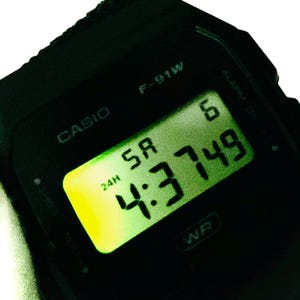 May include: Black Casio F-91W digital watch displaying the time 4:37:49. The watch face has a green digital display with the day of the week and the 24-hour time format. The watch is a classic design.