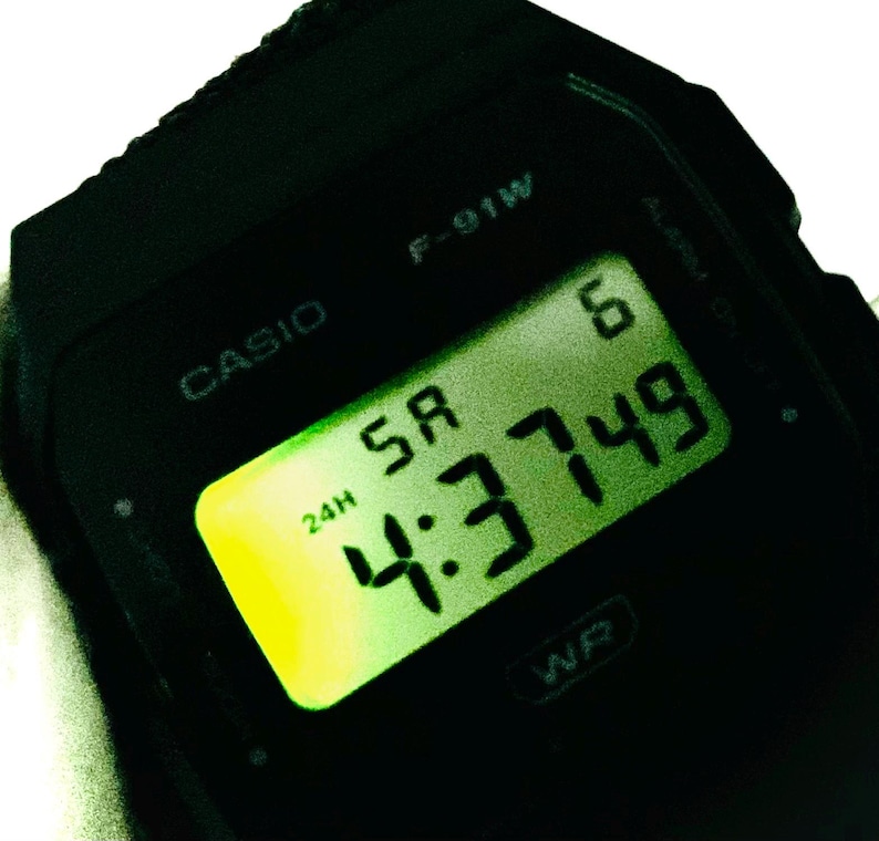 May include: Black Casio F-91W digital watch displaying the time 4:37:49. The watch face has a green digital display with the day of the week and the 24-hour time format. The watch is a classic design.