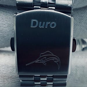 May include: A silver watch strap with a silver clasp. The clasp has the word "Duro" engraved on it and a small marlin fish design.