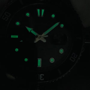 May include: A black watch face with green luminous markings. The watch face has a black background with green luminous hour markers, minute markers, and hands. The watch face also has a green luminous bezel with numbers.