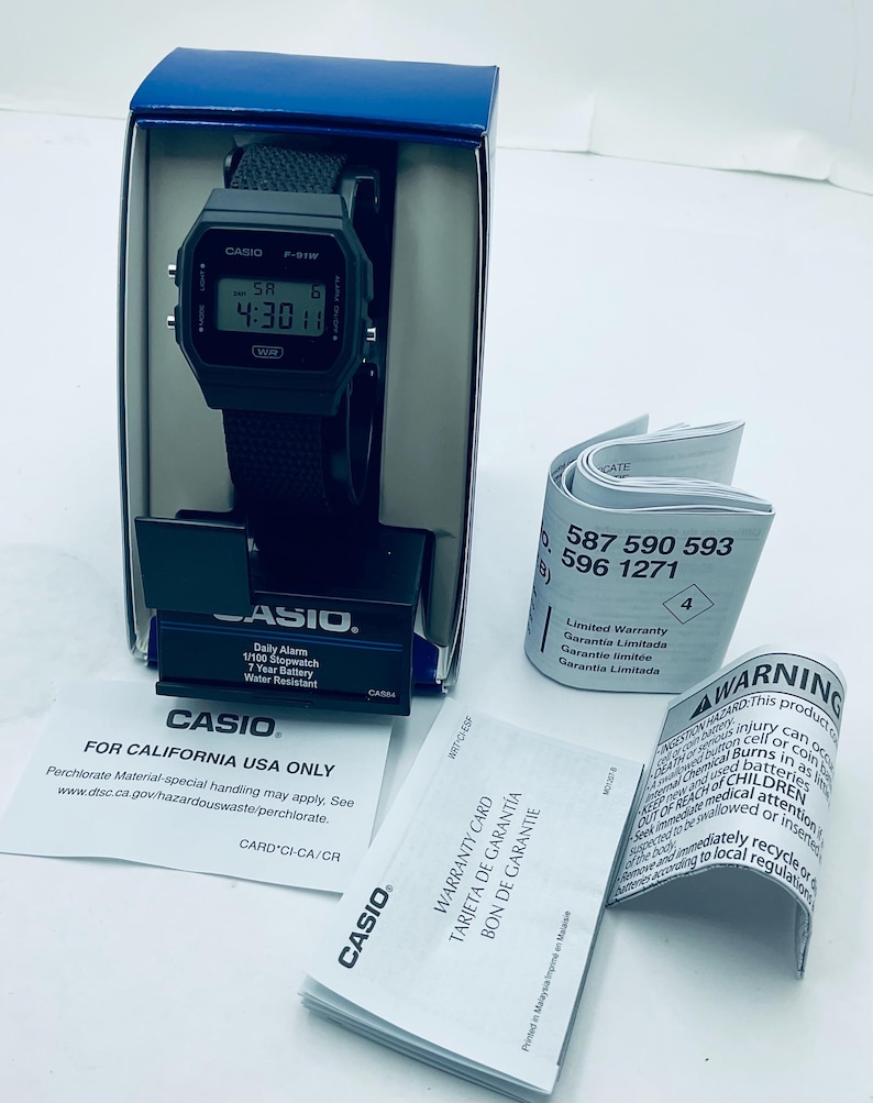 May include: A black Casio F-91W digital watch with a black strap, displayed in its original blue box. The watch face shows the time. Included are warranty cards and a warning label. The watch is water-resistant.