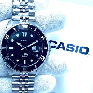 May include: A silver Casio wristwatch with a black face and a silver metal band. The watch has a black bezel with white markings and the words "WR 200M" on the face. The watch is on a white background with the word "CASIO" in blue letters.