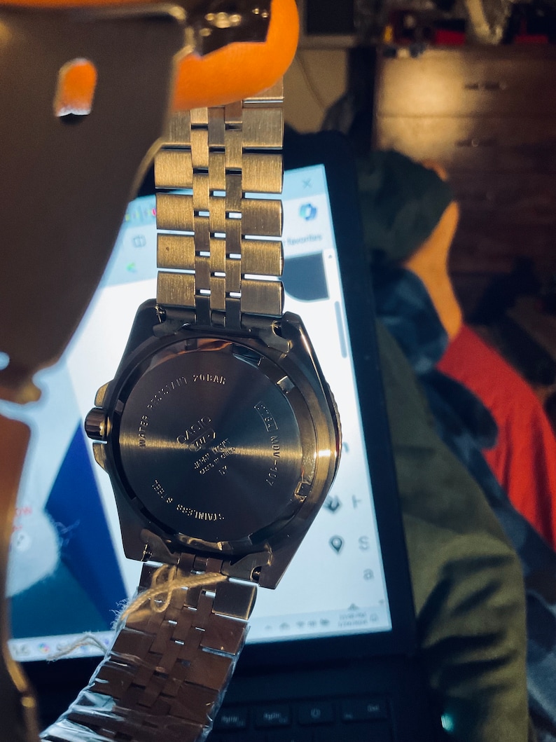 May include: A silver watch with a stainless steel strap. The watch face is black with white markings. The back of the watch has the text "WATER RESIST 20BAR" and "CASIO" printed on it.