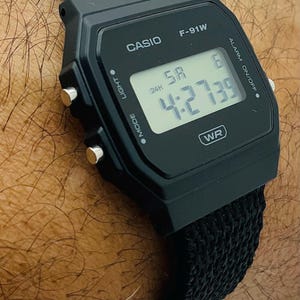 May include: Black Casio F-91W digital watch with a black fabric band. The watch face displays the time, day, and date. The watch has buttons for light, mode, and alarm functions. The time displayed is 4:27:39.
