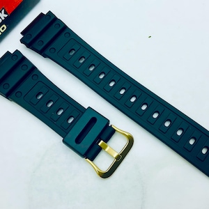 Op de afbeelding: Zwarte vervangende horlogeband met een goudkleurige gesp. De band is gemaakt van een flexibel materiaal en heeft een reeks rechthoekige gaten voor aanpassing. Het G-Shock Casio-logo is zichtbaar op de achtergrond.