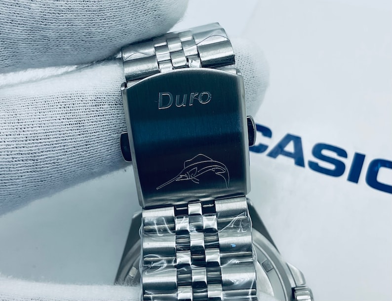 May include: A silver watch with a stainless steel band. The watch has a silver clasp with the word "Duro" engraved on it. A marlin fish is also engraved on the clasp.