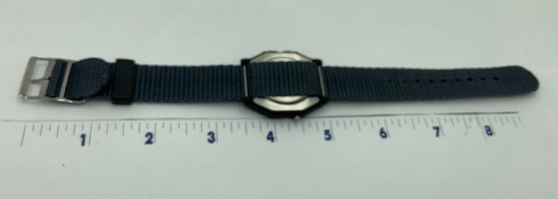 May include: A gray nylon watch strap with a silver watch face. The strap is 8 inches long.