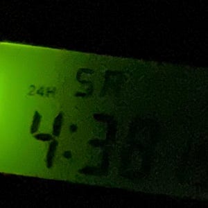 May include: A digital clock with a green illuminated display showing the time as 4:36 AM. The clock face is rectangular with rounded corners, and the background is black.
